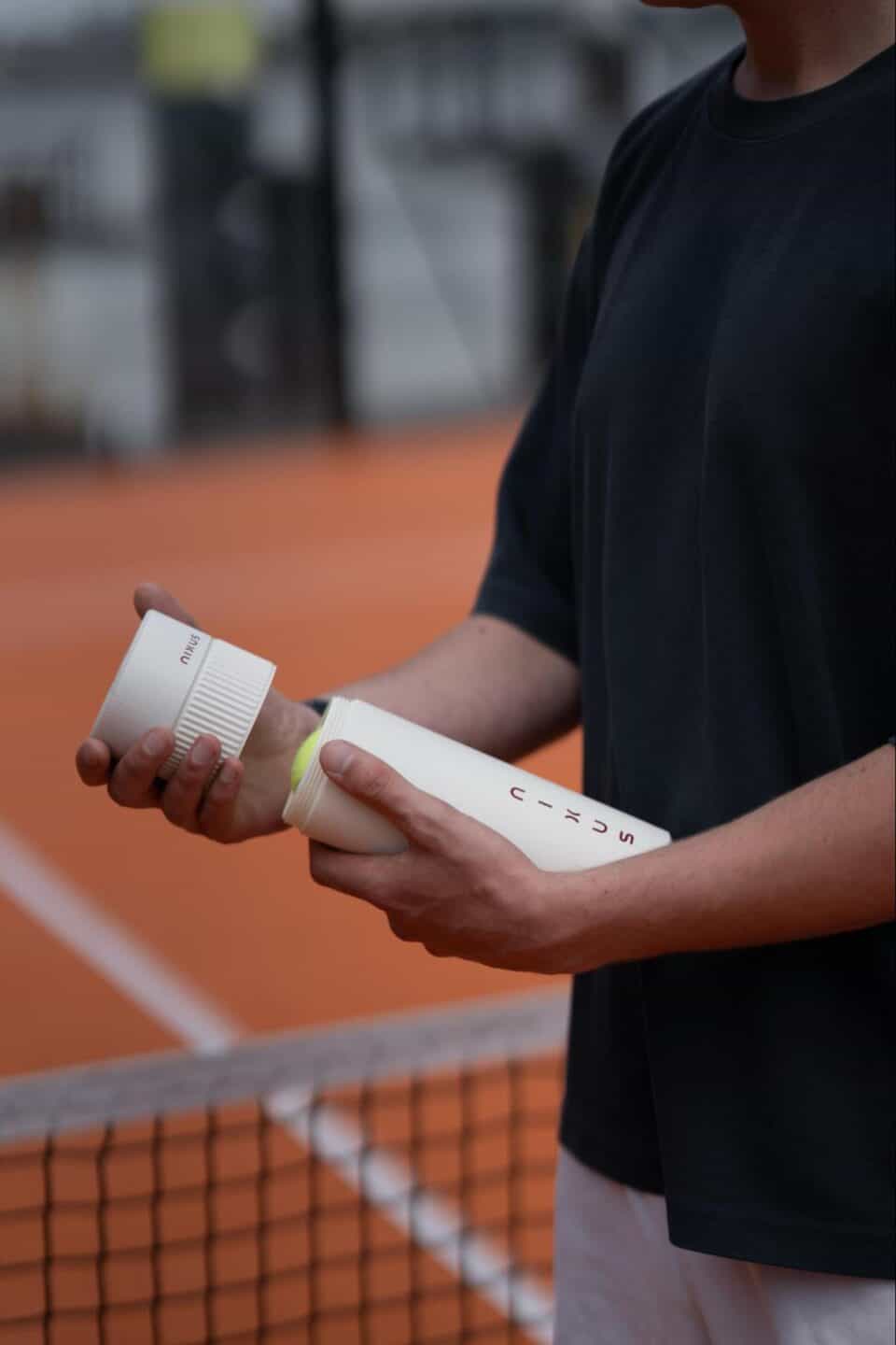 Mid-section view of a person over a tennis net holding an open white NIXUS canister containing a yellow ball.