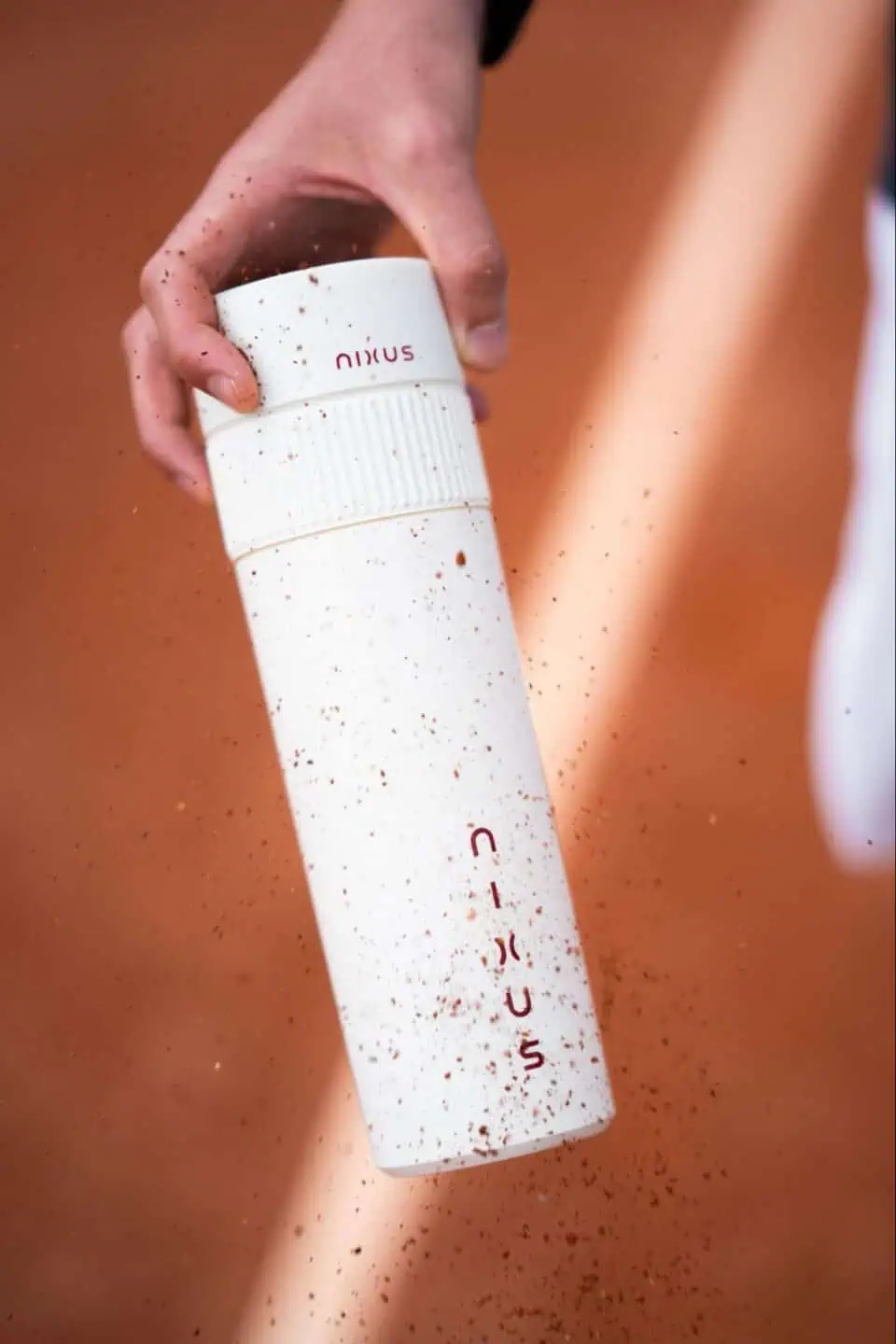 Action shot of a person holding the white NIXUS canister on a tennis court as red clay dust sprays across the frame.